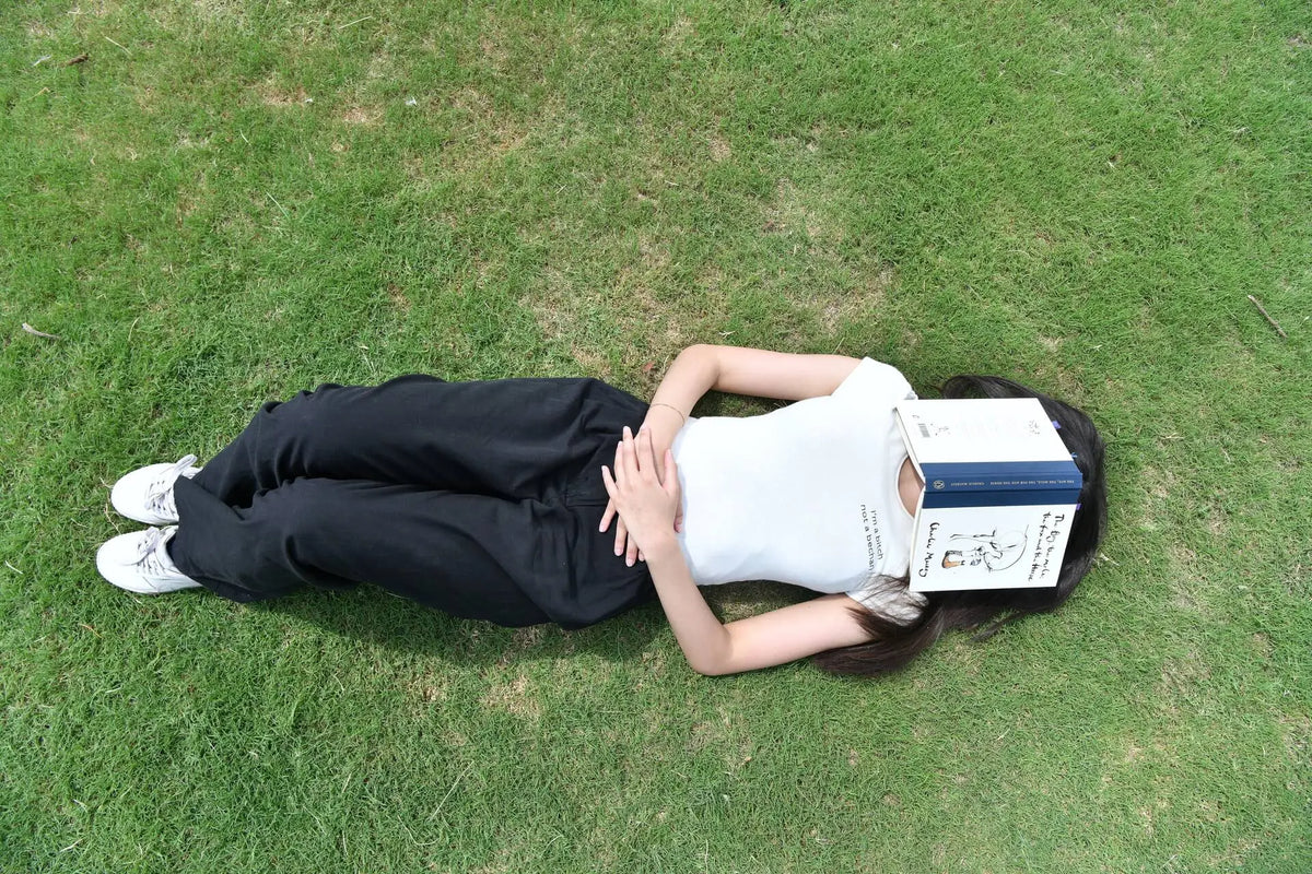 A person lying on green grass, wearing a white sleeveless top and black pants, with a book covering their face.